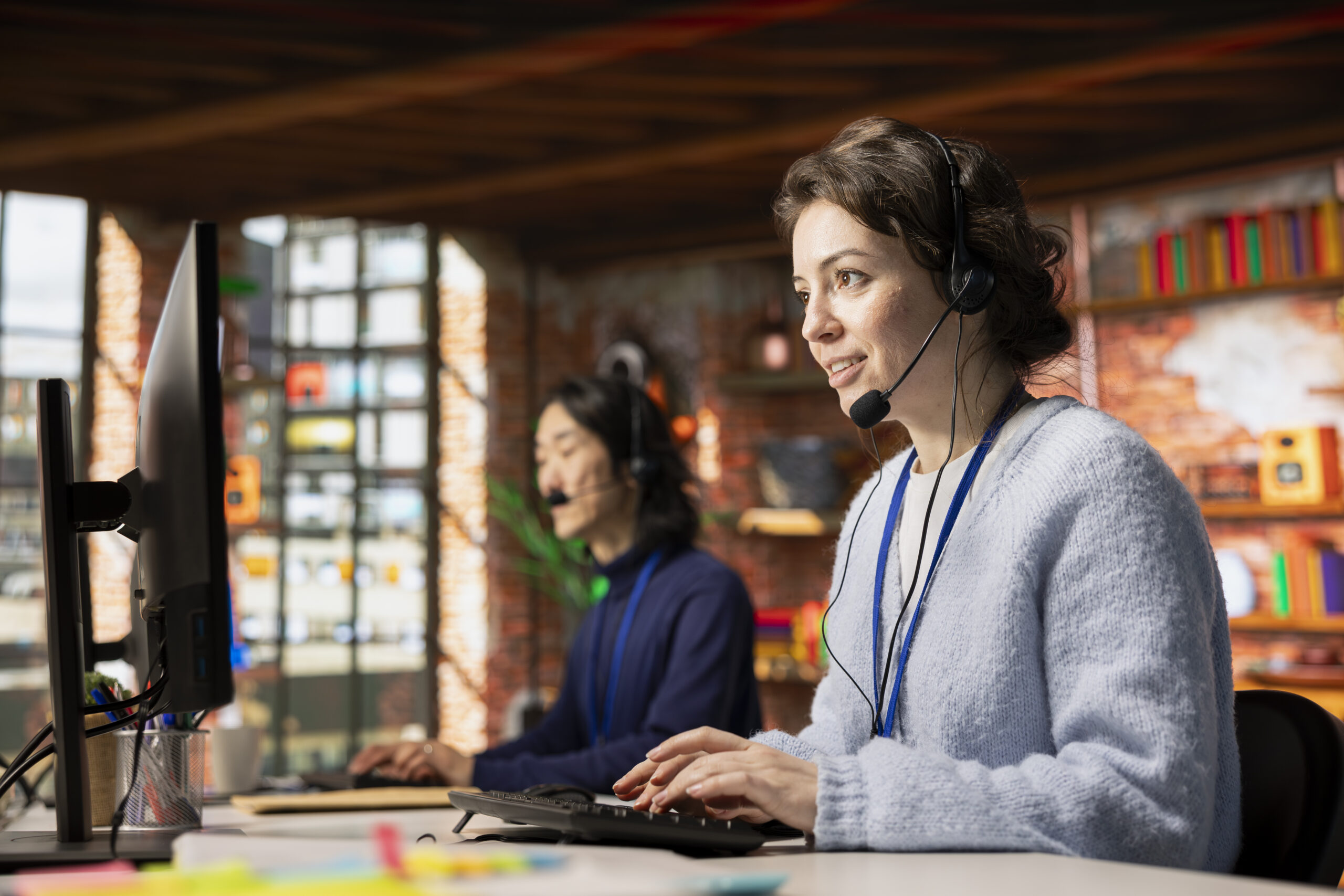 Jolly call center worker dealing with upset customers, speaking using headset at office desk. Friendly woman doing customer support job in workplace using computer, handling clients complaints
